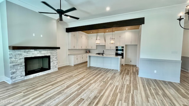 kitchen featuring light wood-type flooring, white cabinets, a stone fireplace, hanging light fixtures, and an island with sink