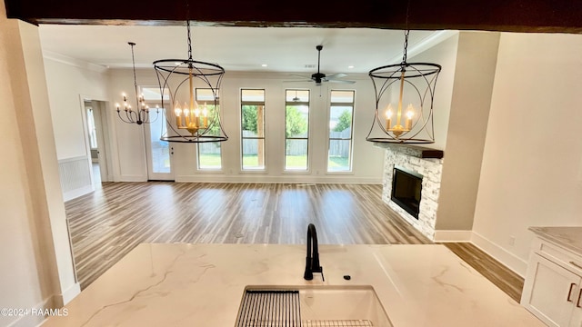 unfurnished living room with sink, a stone fireplace, wood-type flooring, ceiling fan with notable chandelier, and ornamental molding