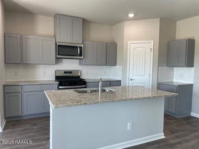 kitchen featuring gray cabinets, a center island with sink, appliances with stainless steel finishes, light stone countertops, and dark hardwood / wood-style flooring