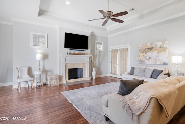 living room with dark wood-type flooring, a tile fireplace, ceiling fan, ornamental molding, and a tray ceiling