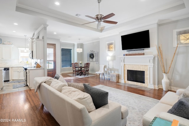 living room featuring ceiling fan, hardwood / wood-style floors, a tray ceiling, a tiled fireplace, and ornamental molding