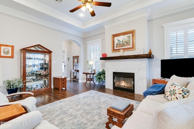 living room with dark wood-type flooring, a tray ceiling, a high end fireplace, crown molding, and ceiling fan