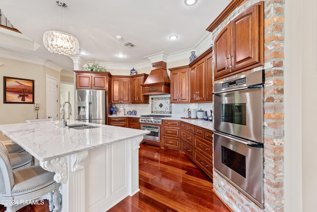 kitchen with premium range hood, appliances with stainless steel finishes, decorative light fixtures, sink, and an island with sink