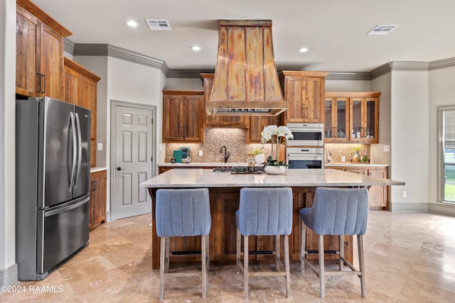 kitchen with a breakfast bar, stainless steel appliances, custom range hood, an island with sink, and decorative backsplash