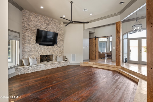 unfurnished living room featuring a stone fireplace, ceiling fan with notable chandelier, crown molding, a barn door, and french doors