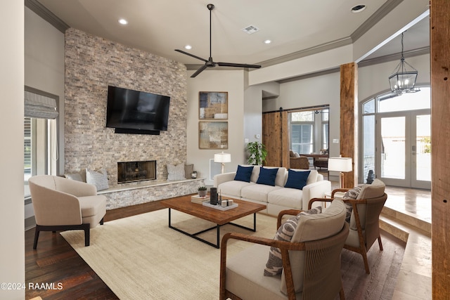 living room featuring a stone fireplace, hardwood / wood-style floors, ceiling fan with notable chandelier, crown molding, and french doors