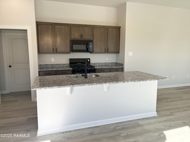 kitchen featuring an island with sink, a kitchen breakfast bar, light stone countertops, dark brown cabinetry, and black appliances