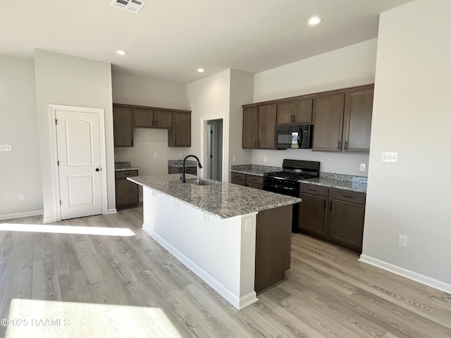 kitchen with black appliances, sink, light wood-type flooring, an island with sink, and stone countertops