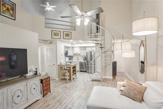 living room featuring light wood finished floors, baseboards, a towering ceiling, ceiling fan, and stairs