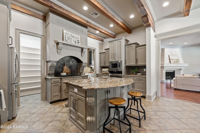 kitchen featuring appliances with stainless steel finishes, a fireplace, a center island with sink, beamed ceiling, and gray cabinets