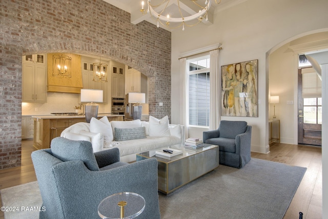 living room with a high ceiling, an inviting chandelier, light hardwood / wood-style flooring, beamed ceiling, and brick wall