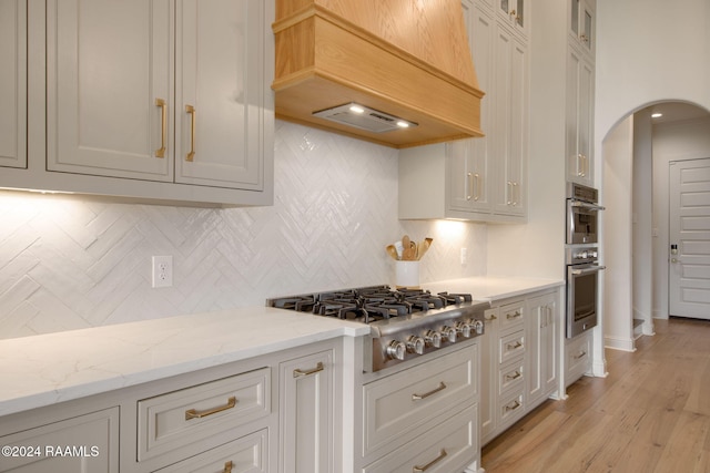 kitchen with light stone countertops, appliances with stainless steel finishes, tasteful backsplash, custom range hood, and light hardwood / wood-style floors