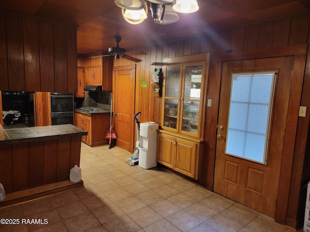 kitchen featuring dobule oven black, under cabinet range hood, stainless steel gas stovetop, wood walls, and brown cabinetry