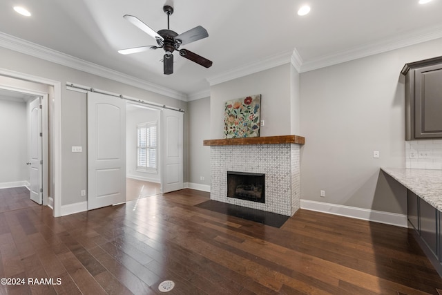 unfurnished living room with a tile fireplace, ceiling fan, a barn door, dark hardwood / wood-style flooring, and ornamental molding