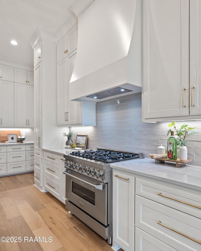 kitchen with premium range hood, light wood-style flooring, high end stainless steel range oven, tasteful backsplash, and white cabinets