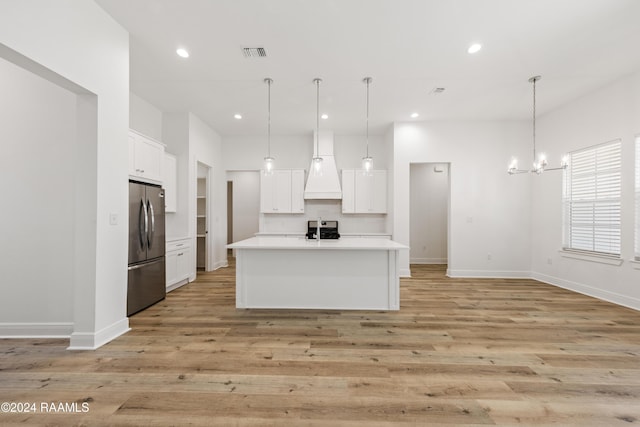 kitchen featuring premium range hood, a center island with sink, stainless steel fridge, decorative light fixtures, and white cabinetry