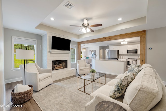 living room featuring a tray ceiling, ceiling fan, sink, a fireplace, and light hardwood / wood-style floors