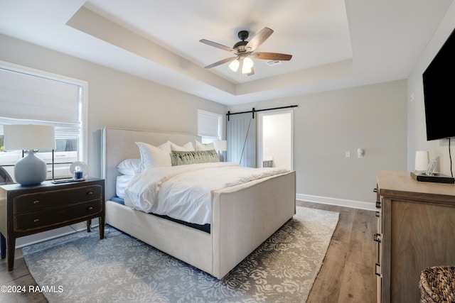 bedroom featuring hardwood / wood-style floors, a barn door, a tray ceiling, and ceiling fan