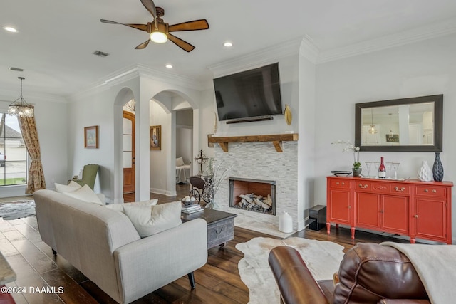 living room featuring dark hardwood / wood-style flooring, a stone fireplace, ceiling fan with notable chandelier, and ornamental molding