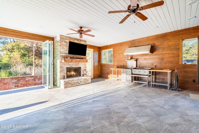 unfurnished living room with a brick fireplace, ceiling fan, wooden ceiling, and wood walls