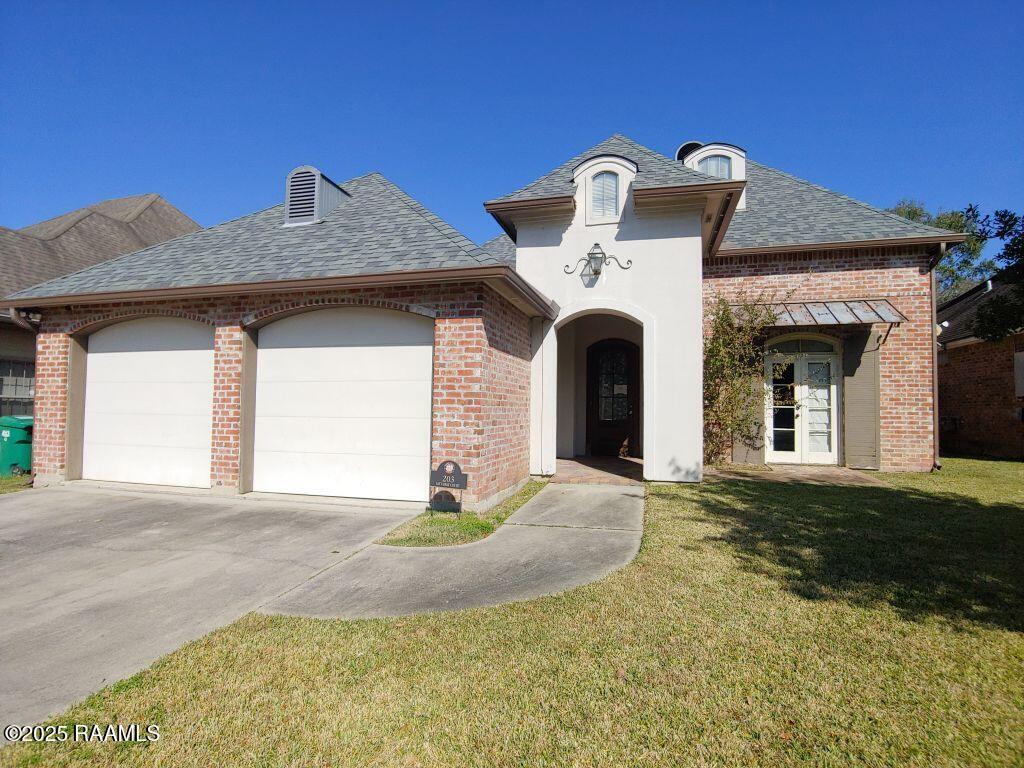 french country style house with a garage, concrete driveway, a front yard, brick siding, and a shingled roof