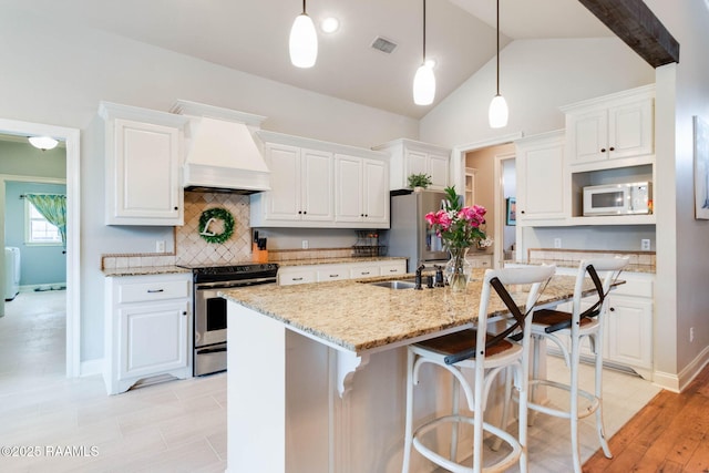 kitchen featuring an island with sink, appliances with stainless steel finishes, custom range hood, decorative light fixtures, and white cabinetry