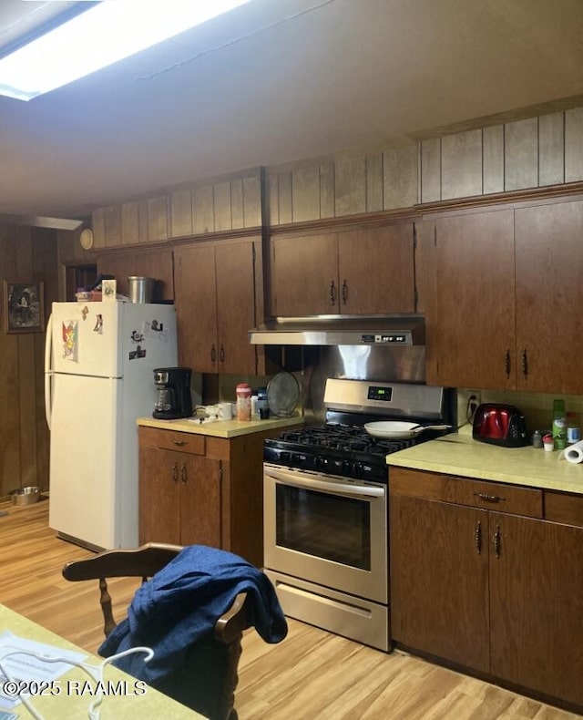 kitchen with wooden walls, light wood-type flooring, white fridge, dark brown cabinetry, and stainless steel range with gas stovetop