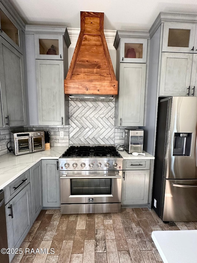 kitchen featuring gray cabinets, appliances with stainless steel finishes, tasteful backsplash, custom exhaust hood, and light stone counters