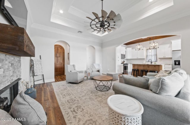 living room featuring a raised ceiling, crown molding, dark hardwood / wood-style flooring, ceiling fan, and a stone fireplace