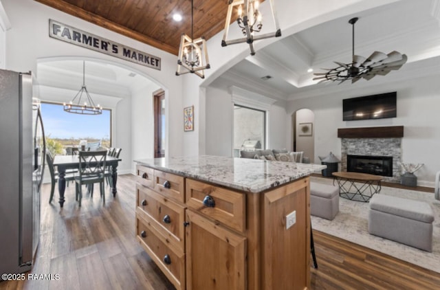 kitchen with hanging light fixtures, stainless steel fridge, a kitchen island, ceiling fan with notable chandelier, and a stone fireplace