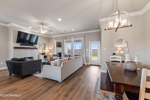 living room with ceiling fan with notable chandelier, a brick fireplace, crown molding, and dark wood-type flooring