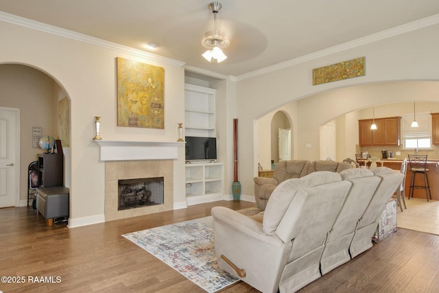 living room with built in features, wood-type flooring, a tiled fireplace, ornamental molding, and ceiling fan