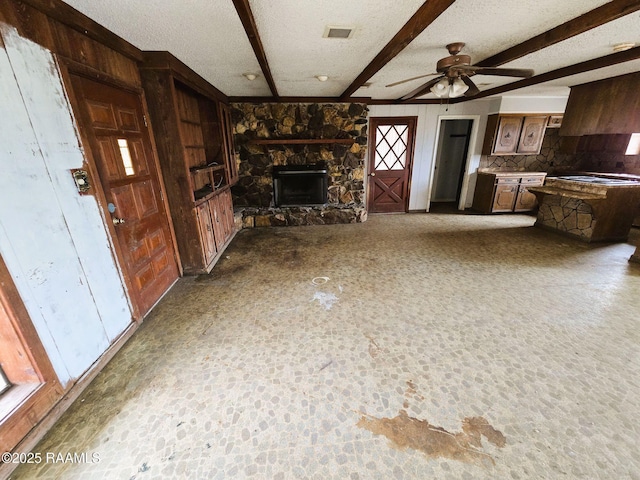 unfurnished living room with beamed ceiling, ceiling fan, a stone fireplace, and a textured ceiling