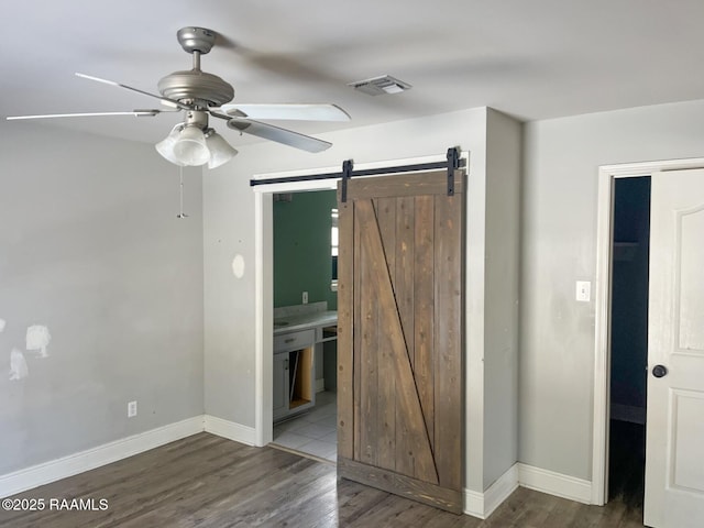 unfurnished bedroom featuring dark wood-type flooring, ceiling fan, ensuite bathroom, and a barn door