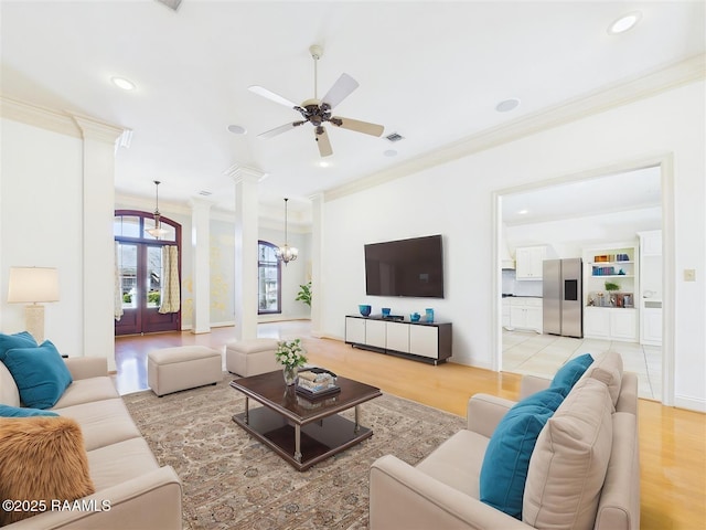 living area featuring light wood finished floors, visible vents, crown molding, decorative columns, and ceiling fan with notable chandelier