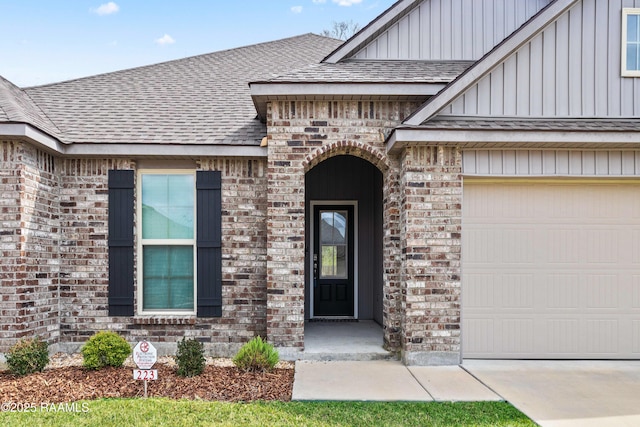 view of exterior entry with an attached garage, brick siding, and roof with shingles