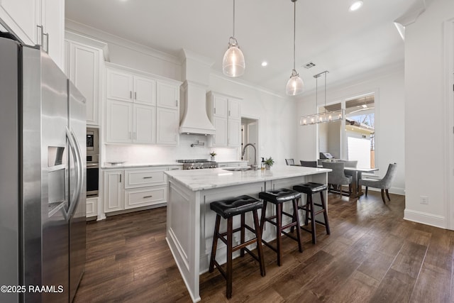 kitchen featuring a center island with sink, custom range hood, pendant lighting, stainless steel appliances, and white cabinets