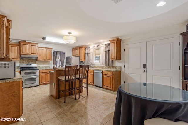 kitchen featuring brown cabinets, under cabinet range hood, appliances with stainless steel finishes, and a kitchen breakfast bar