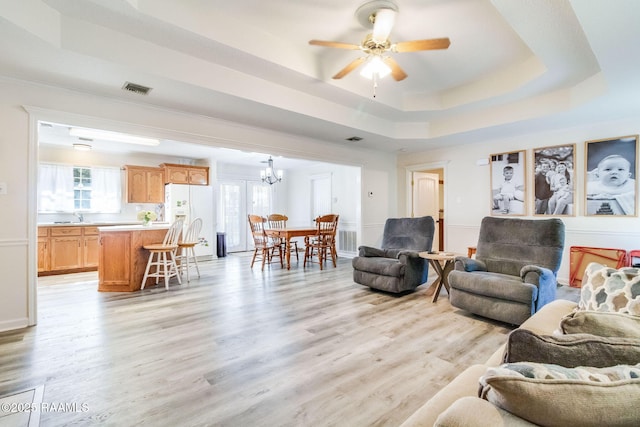 living room featuring sink, a tray ceiling, ceiling fan with notable chandelier, and light wood-type flooring