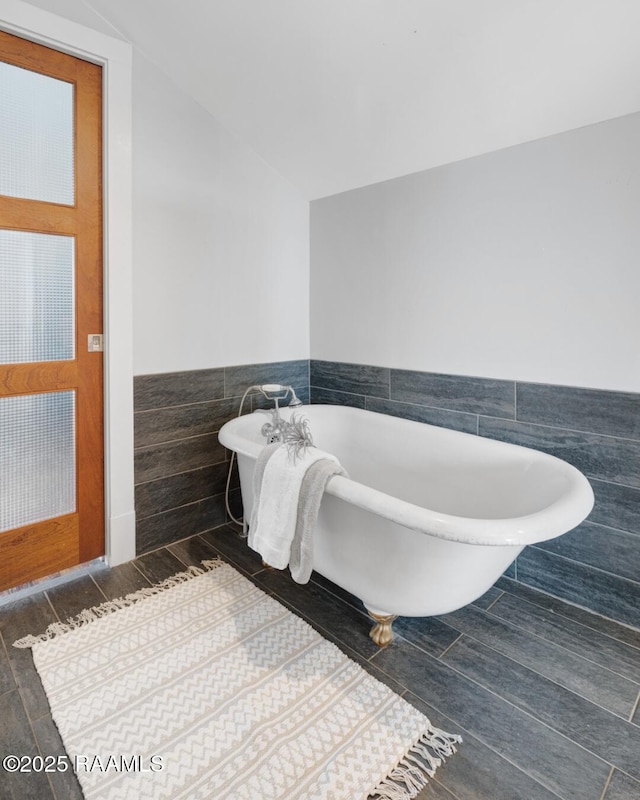 full bath featuring lofted ceiling, a wainscoted wall, a freestanding bath, and tile walls