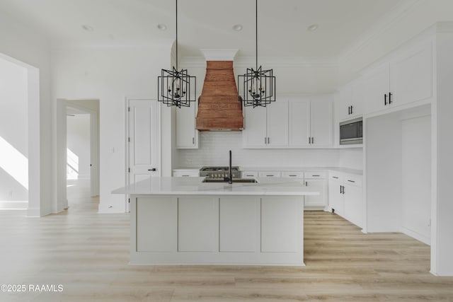 kitchen featuring sink, premium range hood, white cabinetry, a kitchen island with sink, and stainless steel microwave