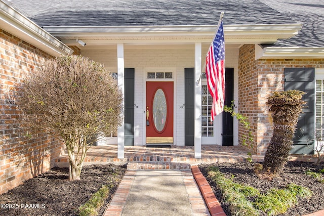 view of exterior entry featuring covered porch, brick siding, and roof with shingles