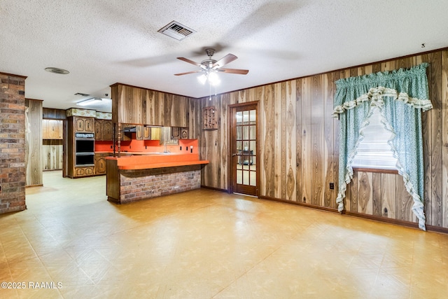 kitchen with black double oven, wood walls, a textured ceiling, ceiling fan, and kitchen peninsula