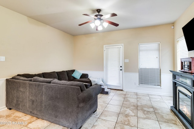 living room with a wainscoted wall, light tile patterned floors, a glass covered fireplace, and a ceiling fan