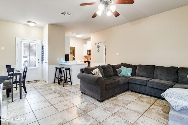 living area featuring wainscoting, light tile patterned flooring, visible vents, and a ceiling fan