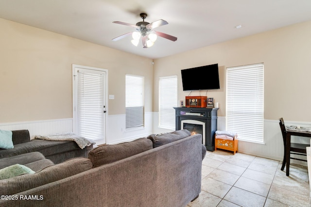 living room featuring a ceiling fan, a glass covered fireplace, wainscoting, and light tile patterned floors