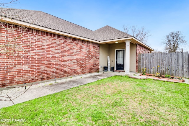 property entrance with a shingled roof, brick siding, fence, and a lawn