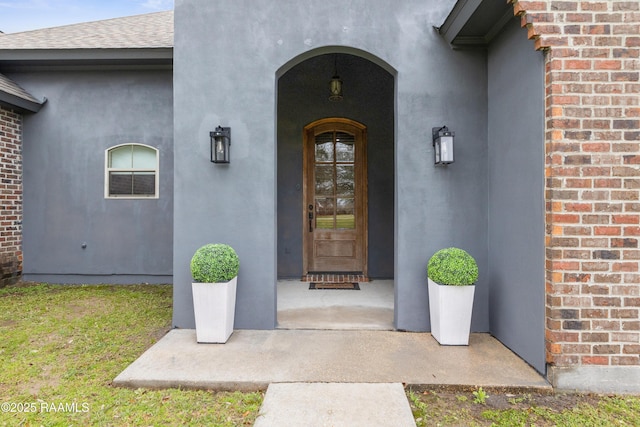 view of exterior entry with a shingled roof, brick siding, and stucco siding