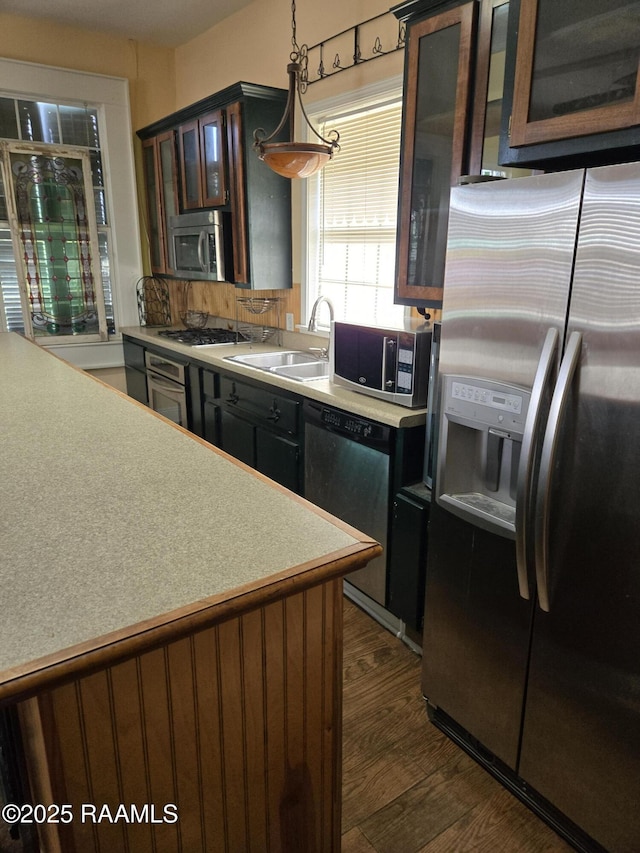 kitchen with a sink, light countertops, appliances with stainless steel finishes, hanging light fixtures, and glass insert cabinets