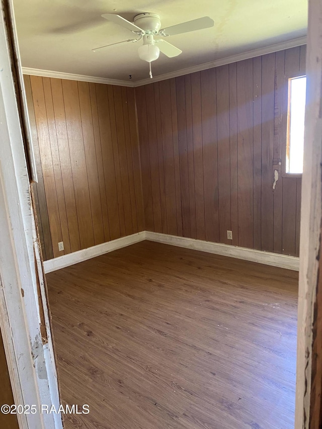 empty room with crown molding, a ceiling fan, wood walls, wood finished floors, and baseboards